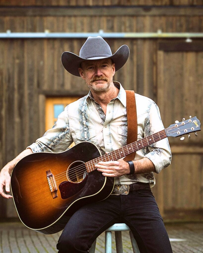 James Bay Market Music for July 2025 - photo of Rob Curtis taken from his facbeook page of him holding his guitar wearing a cowboy hat.