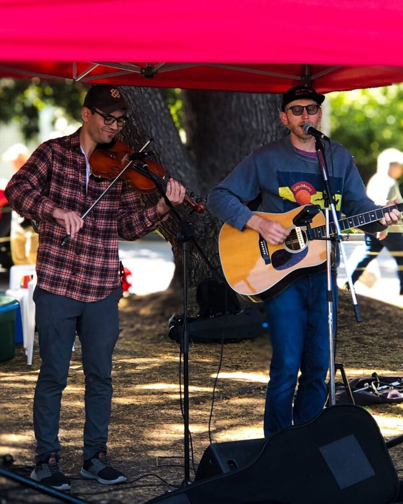 James Bay Market Music for July 2025 - photo is of Tyler James Johnson and his friend playing the fiddle at the JBM.