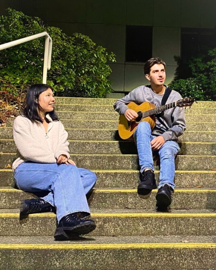 James Bay Market Music line-up for July 2025 - photo is of the duo Jared and Sarah. They are sitting on steps and he is holding his guitar.