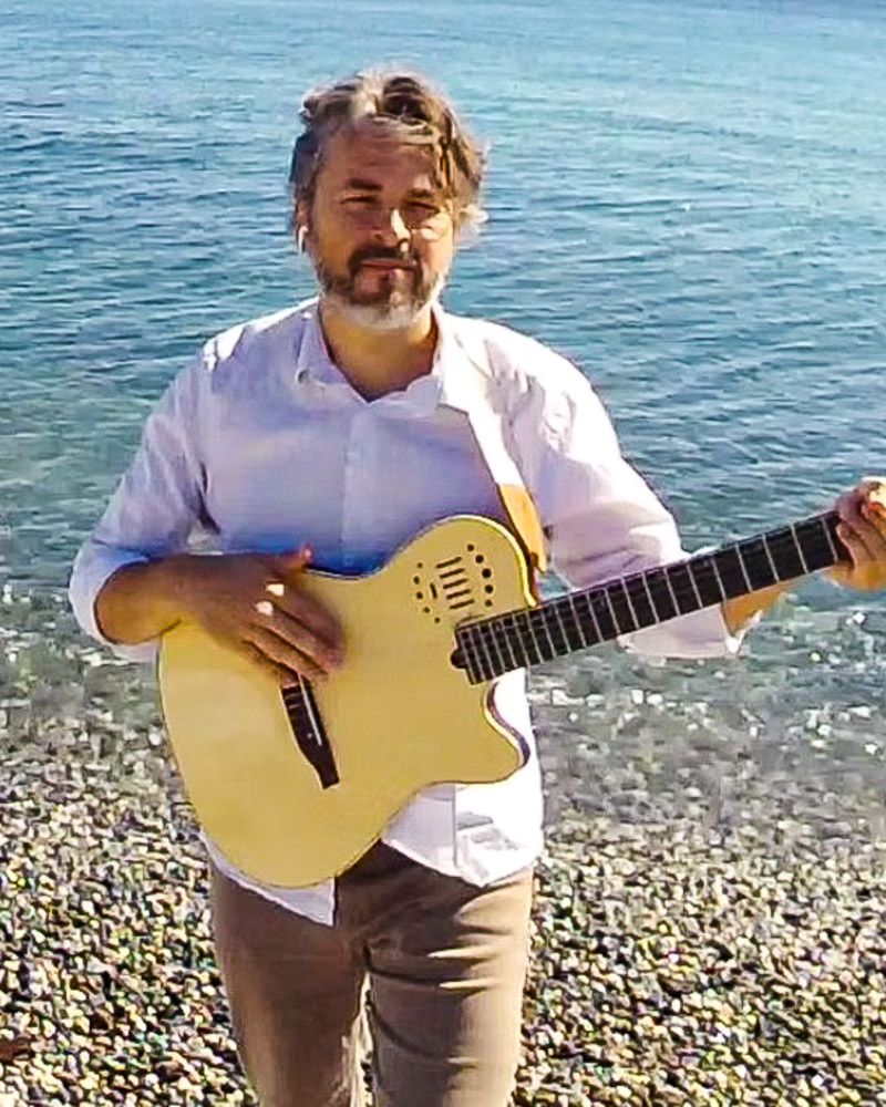 James Bay Market - June 2025 Music Line-up: photo of Dean Wolfe holding his guitar at the beach.